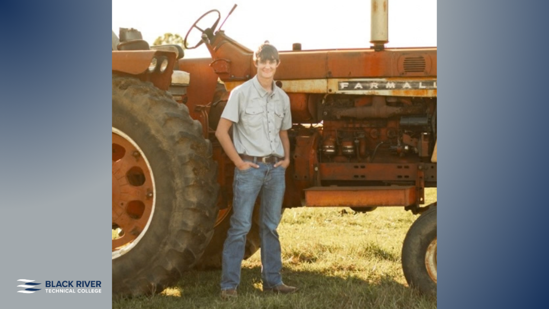 A young man in a gray button-down shirt and jeans stands smiling in front of an old orange Farmall tractor, with Black River Technical College branding.